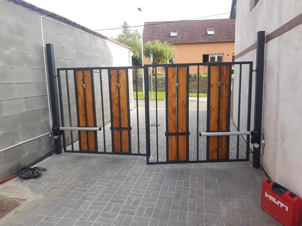 Dvoukřídlá brána z tmavé oceli s výplní z lakovaných dřevěných prken a elektrickým pohonem na dlážděném vjezdu