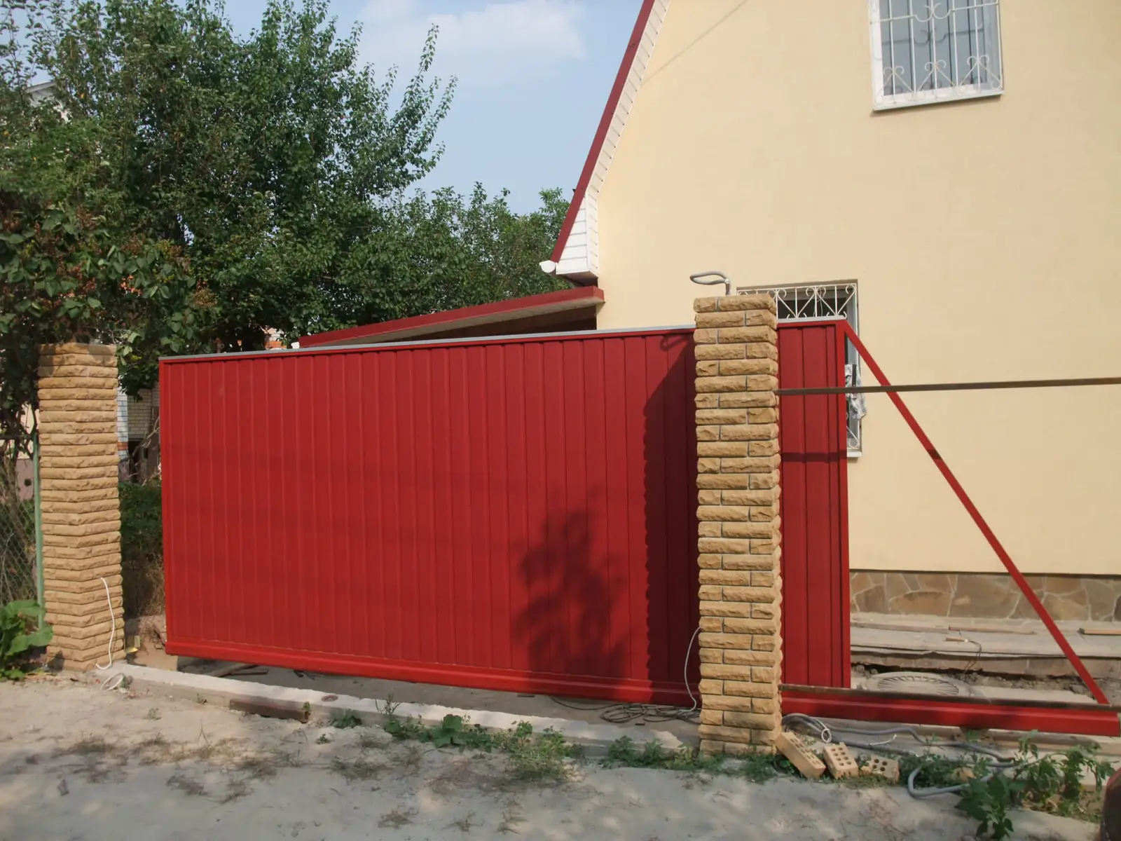 Červená samonosná posuvná brána z trapézového plechu mezi sloupky ze štípaných tvarovek, okolí ještě nedokončené