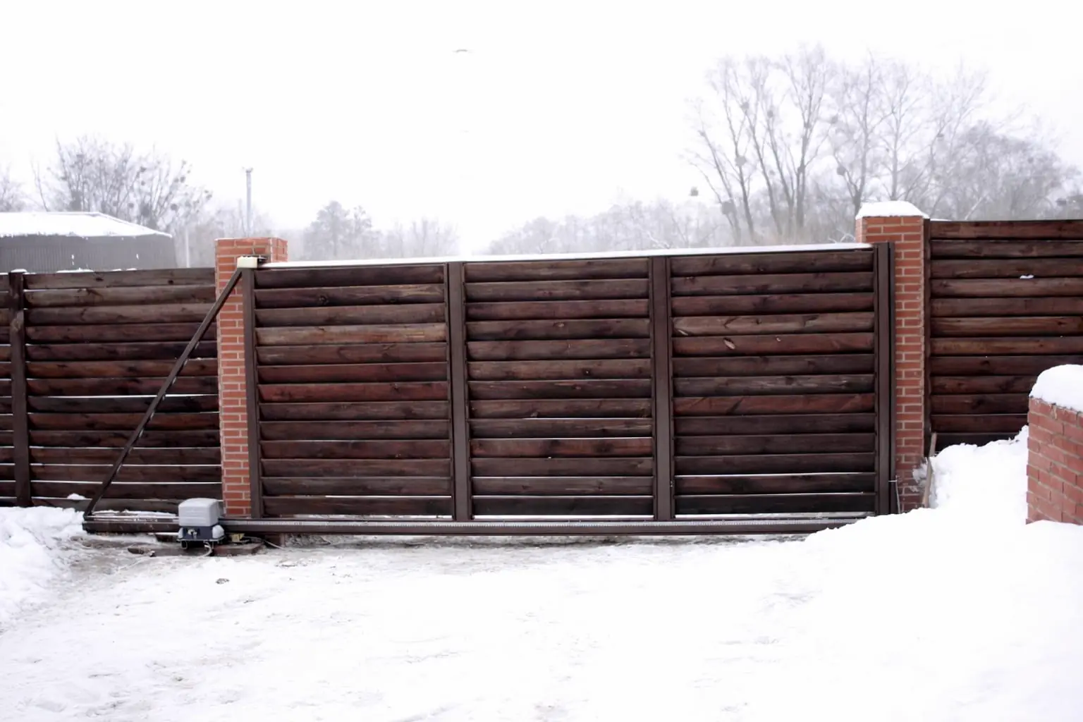 Zasněžený vjezd s posuvnou bránou z hnědých dřevěných lamel v ocelovém rámu mezi sloupky z červených cihel