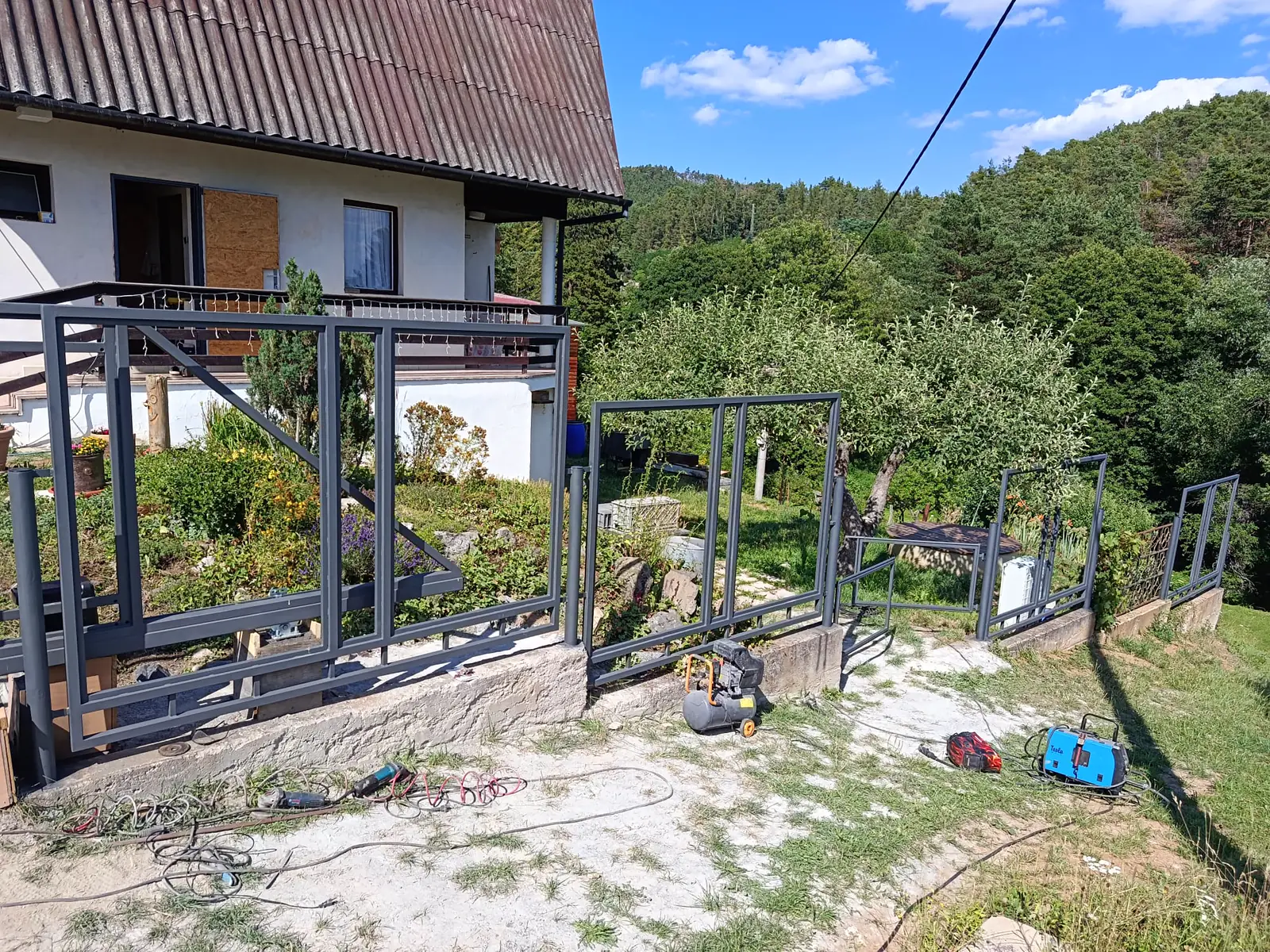 Rozpracovaná posuvná brána a ocelový plot ve svahu u rodinného domu, šedé rámy na betonové podezdívce, v popředí nářadí